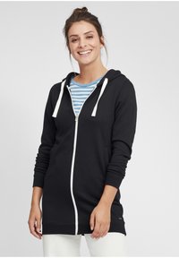 Čierna zip-up mikina s bielymi šnúrkami, dlhými rukávmi, bočnými vreckami a voľným strihom, preložená cez modro-biely pruhovaný tričko.
