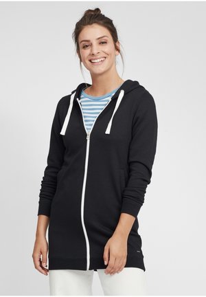 Black zip-up hoodie with white drawstrings, long sleeves, side pockets, and a relaxed fit, layered over a blue and white striped shirt.