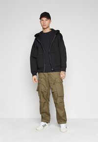 Black zip-up hoodie with large pockets, worn over a dark shirt. Olive green cargo pants with multiple pockets. White sneakers with black stripes.