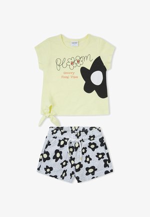 Top amarillo de manga corta con motivo floral en blanco y negro y texto "bloom Groovy Floral Vibes", combinado con pantalones cortos blancos cubiertos de flores negras.