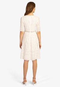 Mujer vistiendo un vestido de encaje blanco hasta la rodilla con mangas tres cuartos y sandalias de tacón blanco, de pie frente a un fondo liso.