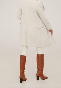 Abrigo de lana beis claro con un corte ajustado, bolsillos laterales y un dobladillo largo, combinado con pantalones blancos ajustados y botas de tacón altas marrones.