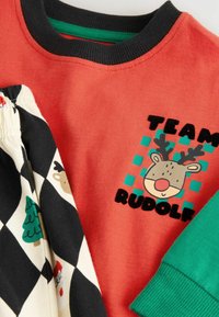 Rotes Baumwollhemd mit einem Rentier-Graphic und dem Text "TEAM RUDOLF". Schwarz-weiße karierte Hose mit Weihnachtsmotiven. Grüne Akzente.