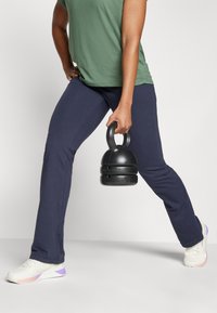 Fekete kettlebell, amit egy kézben tartanak, kopott tengerészkék edzőnadrág, zöld póló és fehér sportcipő lila díszítéssel.