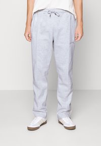 Šedé tepláky vyrobené z měkké tkaniny, s páskem na stažení a diskrétním logem umístěným na levém stehně. Rovný střih nohavic.