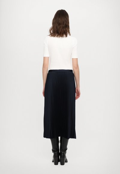 Top bianco a maniche corte abbinato a una gonna midi plissettata scura. La gonna presenta pieghe verticali e una texture liscia, completata da stivali al ginocchio.