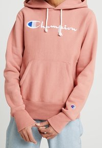 Vaaleanpunainen hupullinen collegepaita pehmeästä kankaasta, jossa on etutasku. Rinnassa ja hihassa valkoinen "Champion"-logo ja sininen yksityiskohta.