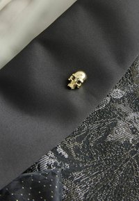 Black satin fabric with a gold skull pin on the lapel, paired with textured black fabric featuring a floral pattern and a dark patterned pocket square.
