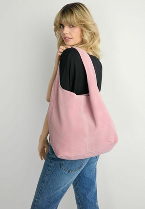 Blondynka w czarnej bluzce i niebieskich dżinsach, niosąca dużą miękką różową torbę na ramię, spoglądająca przez ramię na jednolitym tle.