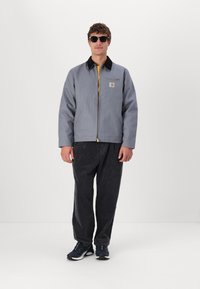 Veste à capuche grise avec un col noir, une fermeture éclair et une poche poitrine. Portée avec un pantalon en velours côtelé foncé et des baskets noires. Design léger et décontracté.
