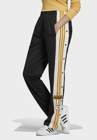Person som bär svarta Adidas träningsbyxor med guld- och vitrandiga detaljer samt tryckknappar, tillsammans med vita Adidas sneakers och en gul topp.