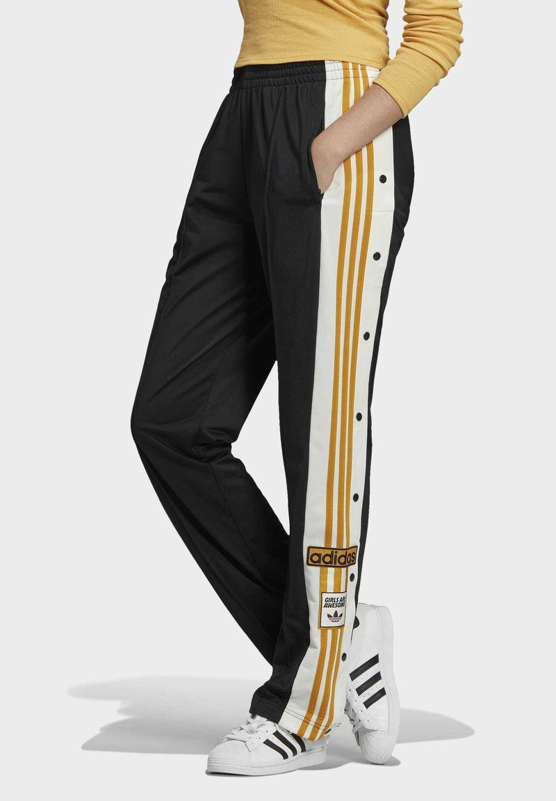 Person som bär svarta Adidas träningsbyxor med guld- och vitrandiga detaljer samt tryckknappar, tillsammans med vita Adidas sneakers och en gul topp.