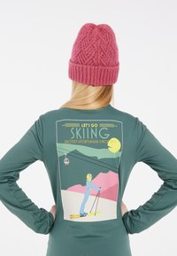 Groen langslaviaan shirt met een skigrafiek op de achterkant met bergen, een skilift en tekst. Roze gebreide muts boven lang haar.