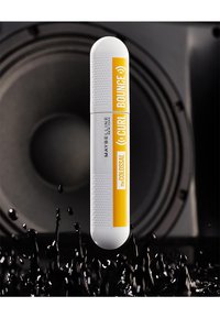 Le tube de mascara Maybelline présente une base blanche avec une étiquette jaune sur laquelle est inscrit "Curl Bounce". Une zone de prise texturée avec un capuchon arrondi. L'arrière-plan montre des ondes sonores.
