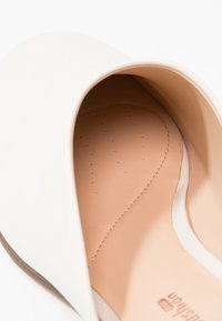 Gros plan de l’intérieur d’une chaussure en cuir beige clair montrant une semelle intérieure rembourrée avec de petites perforations et un détail de couture incurvée.