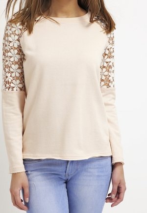 Mujer con una blusa beige de manga larga con paneles de encaje floral en los hombros y jeans azul claro, posando frente a un fondo blanco.