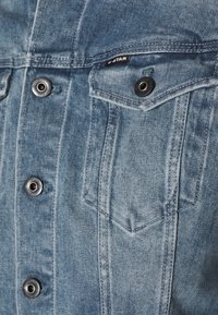 Chaqueta de denim azul claro con botones de presión metálicos, un bolsillo en el pecho con solapa y detalles de costura visibles. La tela tiene una textura suave.