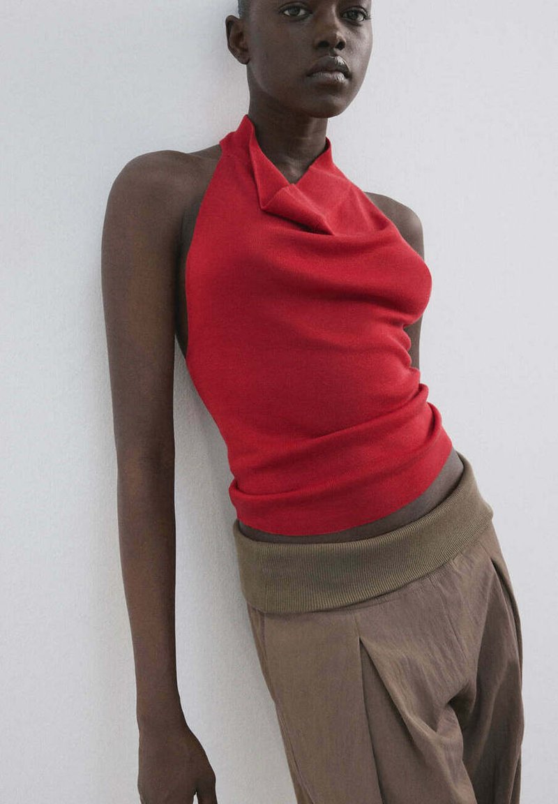 Femme portant un haut dos nu rouge et un pantalon marron taille haute, appuyée contre un mur blanc avec une expression neutre.