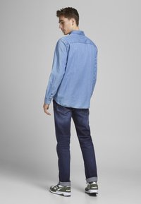 Cămașă din denim albastru deschis cu manșete cu nasturi, asortată cu blugi slim albastru închis și adidași cu accente verzi. Fundal gri neutru.