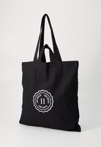 Črn platneni tote nahrbtnik z dvema ročajema in belim logotipom, ki vsebuje krožno zasnovo in besedilo. Gladka tekstura, pravokotna oblika.