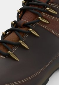 Bota de senderismo de cuero marrón con acentos marrones texturizados, cordones negros y ojetes metálicos dorados. Presenta un contraste de costuras blancas.