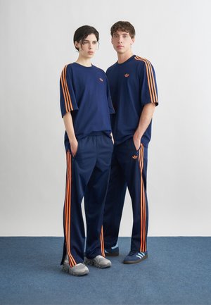 Δύο άτομα στέκονται δίπλα δίπλα φορώντας σκούρα μπλε φόρμες με πορτοκαλί ρίγες και λογότυπα, με τα χέρια στις τσέπες και ουδέτερες εκφράσεις.