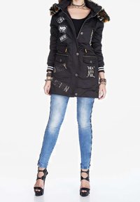 Parka nera con cappuccio foderato in pelliccia, caratterizzata da toppe e testo ricamato con paillettes. Abbinata a jeans skinny azzurri e tacchi neri con cinturini.