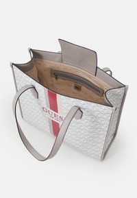 Sac fourre-tout en matière blanche à motifs avec des rayures rouges et beiges. Contient une doublure beige et une poche zippée. Dispose de poignées grises.