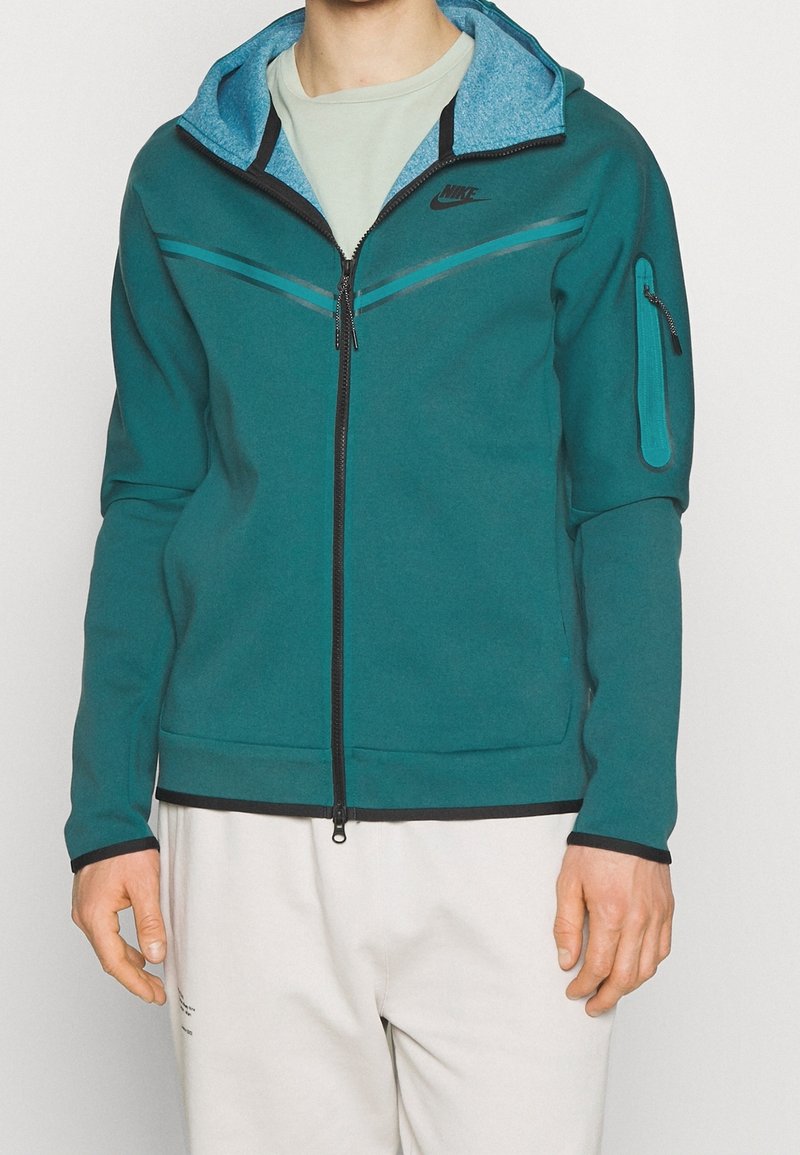 Man i en teal zip-up Nike-jacka med huva och dragkedjefodral på ärmen, tillsammans med ljusa byxor.