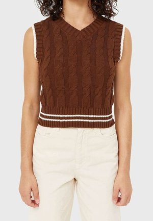 Personne portant un gilet sans manches en maille torsadée marron avec des rayures blanches sur une chemise blanche et un pantalon clair.
