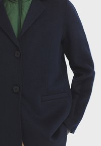Abrigo de mezcla de lana en color marino con solapa, dos botones en la parte delantera y un bolsillo lateral. Textura suave con una silueta limpia y entallada.