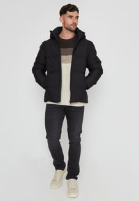 Sort polstret jakke med hætte, lynlåslukning og sidelommer. Båret over en flerfarvet stribet sweater, parret med mørke jeans og lyse sneakers.