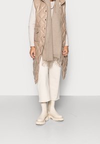 Kvinna iförd beige vadderad lång väst, beige scarf, krämfärgad tröja, vida krämfärgade byxor och krämfärgade anklar med tjock sula mot en enfärgad bakgrund.