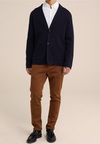 Modrý pletený kardigan s šálovým límcem, dvěma předními kapsami a zapínáním na jeden knoflík, doplněný o hnědé úzké kalhoty a černé boty.