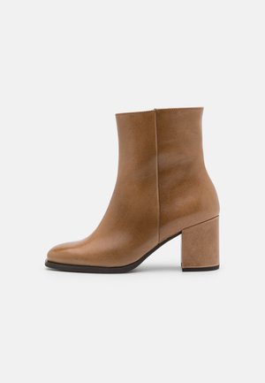 Bottine en cuir couleur tan avec talon bloc, bout arrondi et fermeture éclair latérale sur fond uni gris clair.