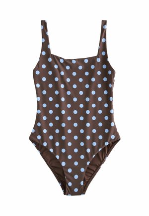 Młoda kobieta o długich ciemnych włosach, nosząca czarne okulary przeciwsłoneczne i brązowy kostium kąpielowy w kropki, stoi na słonecznej plaży.