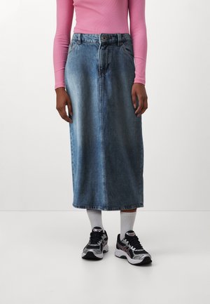 Personne portant une jupe midi en denim bleu délavé, un haut rose à manches longues, des chaussettes blanches et des chaussures de sport noires et grises, debout sur un fond blanc.