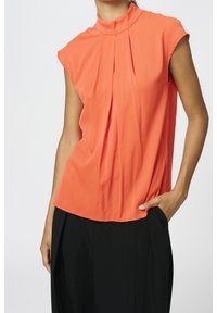 Blouse orange légère avec un col haut, des détails plissés et des manches courtes, associée à un pantalon noir ample.