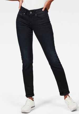 Pantalones vaqueros de mezclilla negra con un diseño ajustado, que cuentan con una cintura alta, cinco bolsillos y un cierre de botón. Combinados con zapatillas blancas.