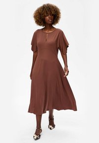 Vrouw met krullend haar in een bruine midi-jurk met vloeiende mouwen en een keyhole-halslijn, gecombineerd met animal print enkellaarsjes met enkelband.