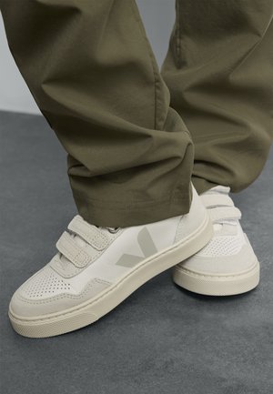 Sneaker bianco con accenti grigi, caratterizzato da una superficie testurizzata, perforazioni e cinghie in velcro. Abbinato a pantaloni verde oliva.