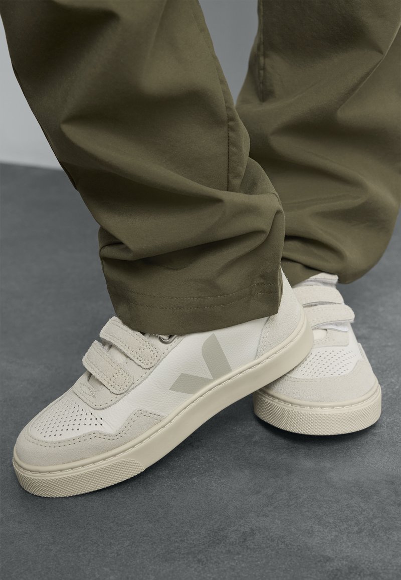Hvite sneakers med grå detaljer, med en strukturert overflate, perforeringer og borrelåsremmer. Kombinert med olivengrønne bukser.