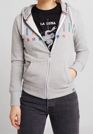 Osoba nosząca szarą bluzę z kapturem na zamek błyskawiczny z kolorowymi literami i symbolami, pod którą ma czarną koszulkę z grafiką "La Luna" z białym wężem.