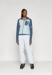 Lichtblauw en marineblauw jack met rits en een zak; gecombineerd met een witte broek en zwarte sneeuwlaarzen. Voorzien van een logo op de mouw.