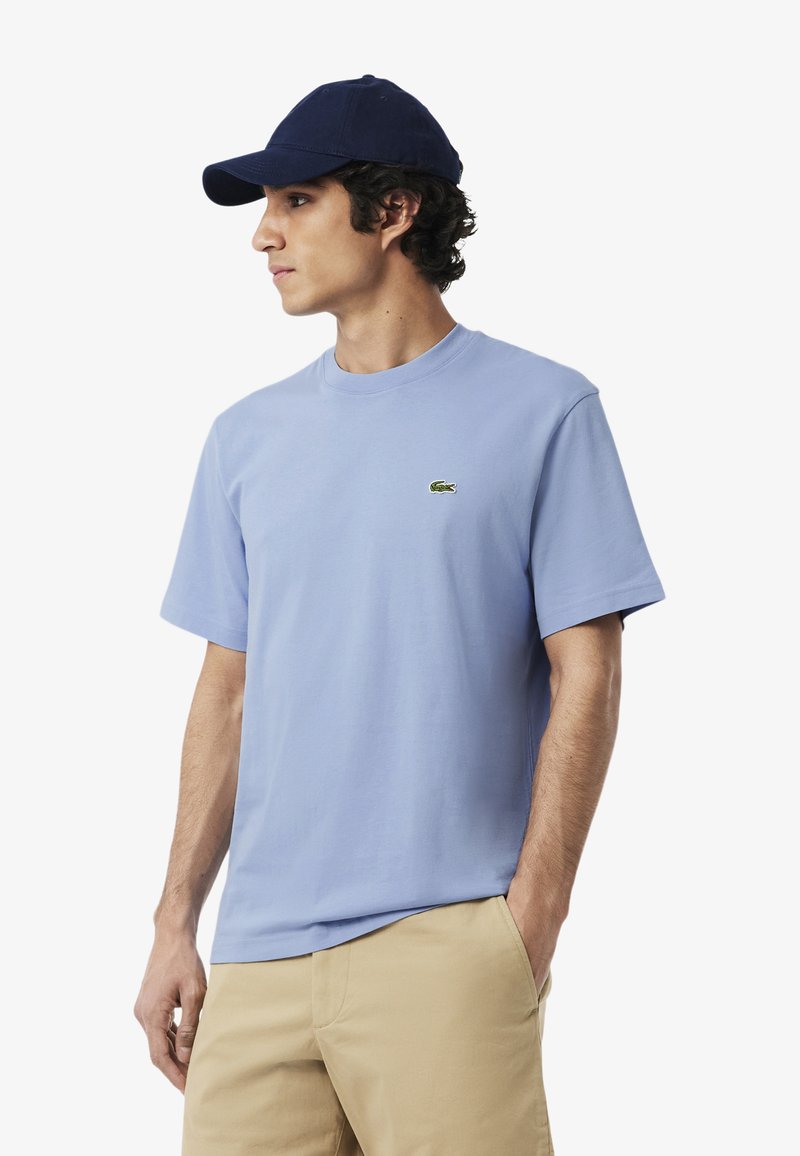 Ung mand iført en ensfarvet lyseblå t-shirt med en lille grøn krokodille-logo og en marineblå kasket, med den ene hånd i lommen på khakibukser.