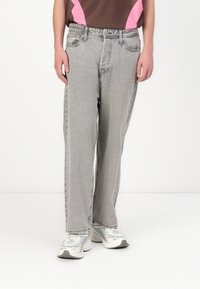 Person klädd i ljusgrå raka jeans och vita sneakers med blå detaljer, stående framför en enfärgad vit bakgrund.