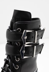 Bota de cuero negro con dos hebillas plateadas, cordones contrastantes con patrones en blanco y negro, y un puño texturizado.