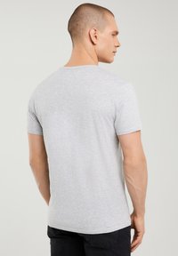 Mand med kort hår iført en ensfarvet lysegrå t-shirt med korte ærmer og sorte bukser, stående med ryggen delvist vendt.