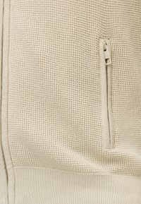 Tissu tricoté beige avec texture gaufrée et ourlet côtelé, avec une poche verticale zippée beige assortie.