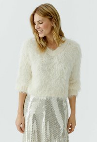 Vrouw die glimlacht, draagt een pluizige witte trui met driekwart mouwen en een glanzende zilveren paillettenrok, tegen een effen witte achtergrond.